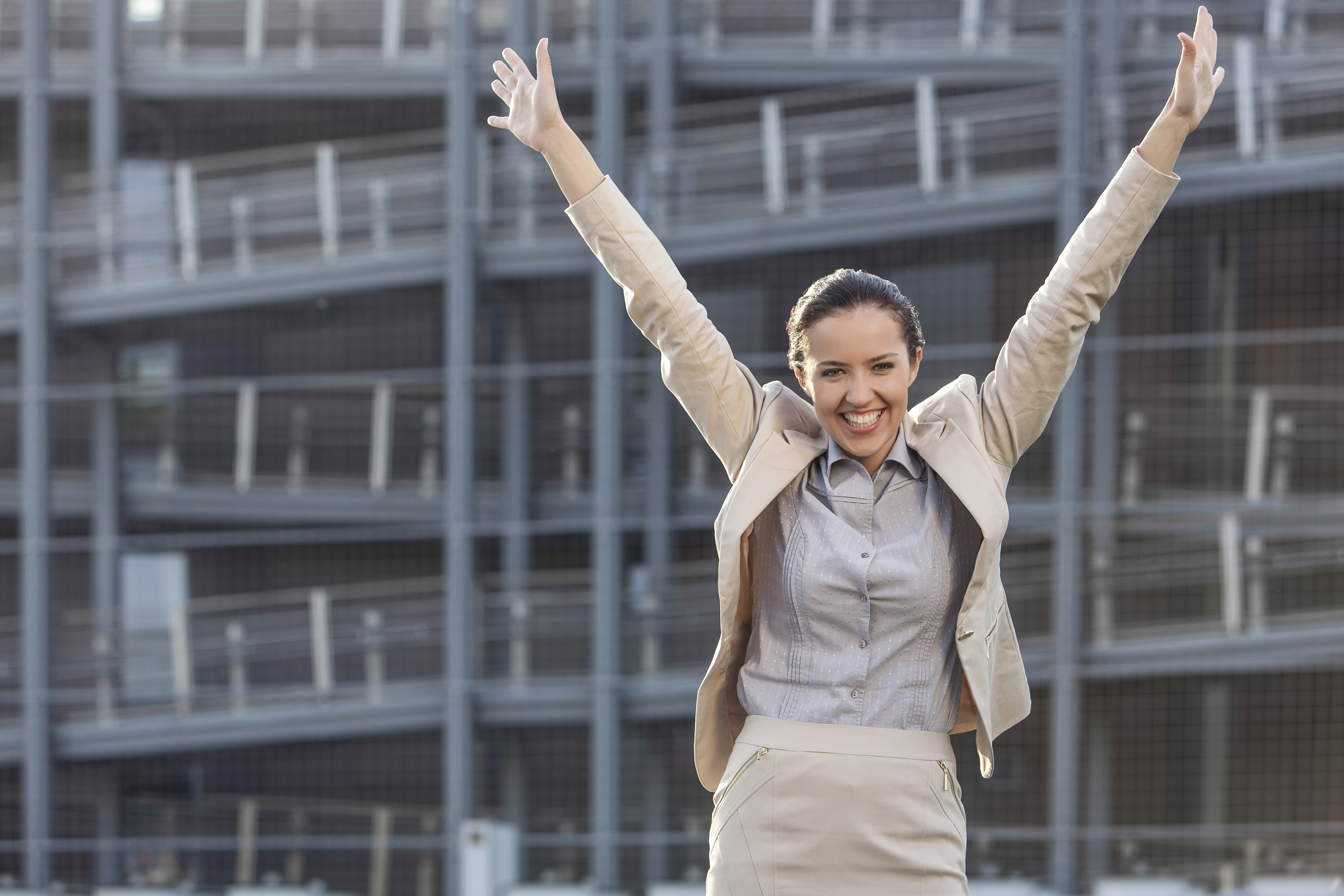 Excited young businesswoman with arms raised standing against office building
